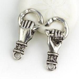 May include: Two silver-toned metal charms shaped like a hand clenching a curved bar. The hand has detailed fingers and is attached to a decorative cuff. Each charm has a small loop at the bottom.