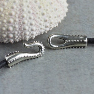 May include: Two silver metal clasps with a textured surface and a hook design. The clasps are attached to a black leather cord.