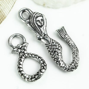 May include: Two silver mermaid charms. One charm features a mermaid with a tail and scales. The other charm features a mermaid with a tail and scales, and a face.