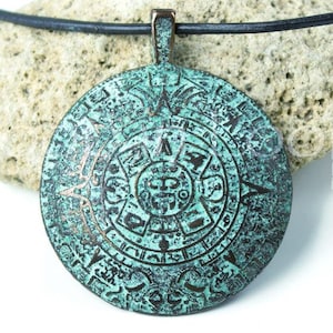 May include: A round, teal and bronze-toned pendant featuring an intricate, embossed circular calendar design. The pendant is suspended from a simple black cord, showcasing a weathered, metallic finish with detailed geometric and symbolic carvings.