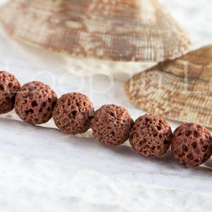 May include: A strand of brown lava rock beads, each bead is round and has a porous texture. The beads are arranged in a line on a white surface.