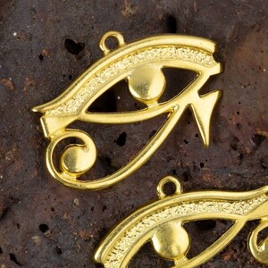 May include: Two gold-colored metal charms in the shape of the Eye of Horus, an ancient Egyptian symbol of protection. The charms are detailed with intricate lines and a raised circle in the center of each eye.