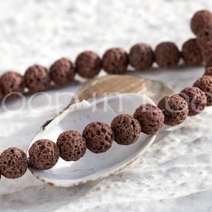 Könnte beinhalten: Eine Kette aus dunkelbraunen Lavasteinperlen, jede mit einer porösen Textur, präsentiert auf einer weißen Muschelschale. Die runden Perlen sind gleichmäßig groß und eignen sich für die Schmuckherstellung oder Bastelarbeiten. Der Hintergrund ist eine strukturierte weiße Oberfläche.