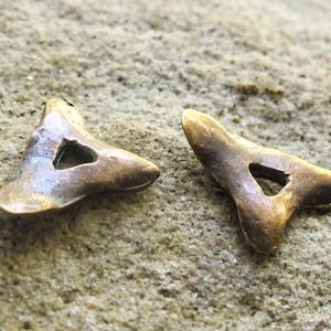May include: Two small, metallic, bronze-colored charms shaped like rounded triangles. Each charm features a smooth, slightly weathered surface with a central triangular cutout, creating a hollow, open-center design with soft, curved edges.