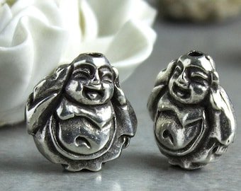 May include: Two silver-colored Buddha beads. Each bead depicts a smiling Buddha with hands near the head. The beads have a detailed, textured surface. The background includes a white flower and a light-colored surface.