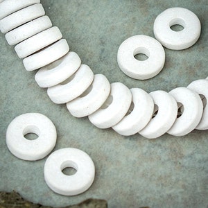May include: A string of white ceramic donut-shaped beads. The beads are arranged in a circle and are all the same size. There are also several loose beads on the surface.