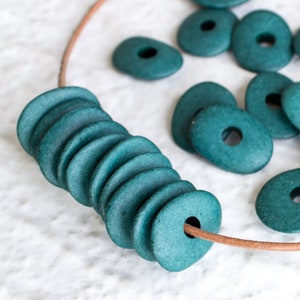 May include: A close-up of a teal green necklace made of stacked ceramic beads with a leather cord. The beads are oval shaped with a hole in the center. There are additional loose beads scattered around the necklace.