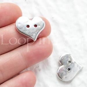 May include: Two silver heart-shaped buttons with two holes each. The buttons are shown on a hand.