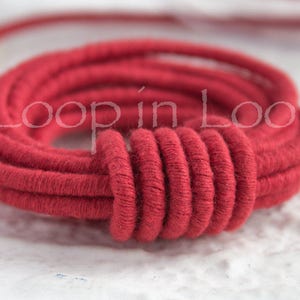 May include: Close-up of a red cotton cord, twisted and coiled into a circle. The cord is thick and soft, with a slightly textured surface.