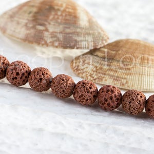 May include: A strand of brown lava rock beads, with a rough, porous texture. The beads are arranged in a line on a white surface, with two seashells in the background.