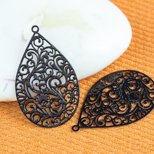 May include: Two black metal filigree teardrop-shaped pendants with intricate designs. The pendants are on a textured orange surface.