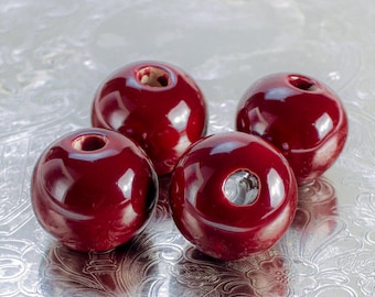 May include: Four round, glossy, dark red ceramic beads with a hole in the centre. The beads are arranged on a silver surface.