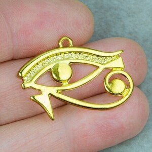May include: Gold-colored metal pendant charm in the shape of the Eye of Horus, an ancient Egyptian symbol of protection and good health.