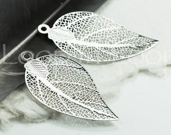 Op de afbeelding: Twee zilveren blaadjes met een delicaat, ingewikkeld ontwerp. De blaadjes hebben een gaatje bovenaan om aan sieraden of andere knutselwerkjes te bevestigen.