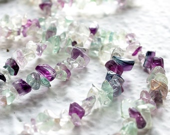May include: A strand of multi-colored fluorite chip beads. The beads are a mix of light green, purple, and clear. The beads are strung on a clear thread.