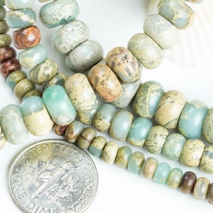 May include: A close-up of a strand of light blue, green, and brown jasper beads. The beads are various sizes and shapes, and some have a marbled pattern. A UK ten pence coin is included for scale.