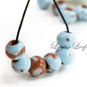 May include: A close-up of a necklace made with light blue and brown ceramic beads. The beads have a speckled pattern and are strung on a black cord.