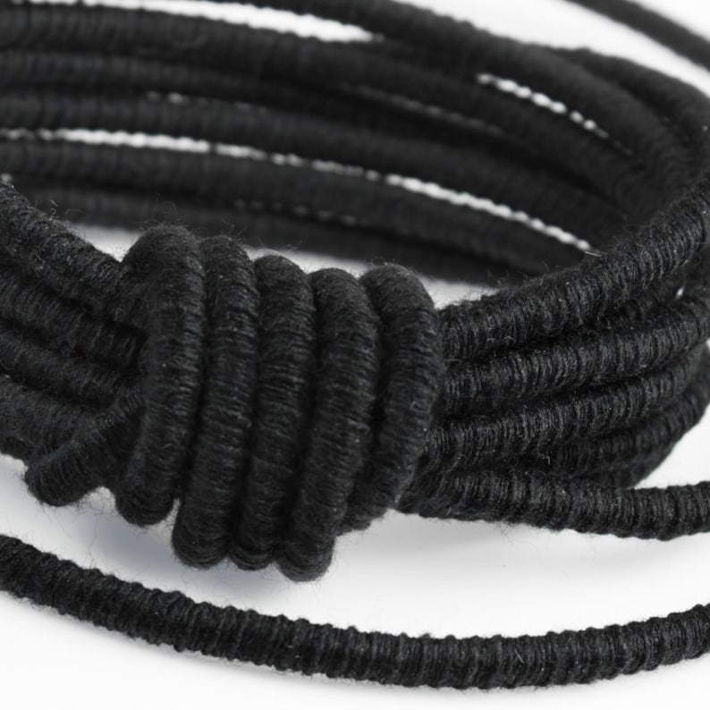May include: A black braided bracelet with a knotted design. The bracelet is made of multiple strands of braided cord.