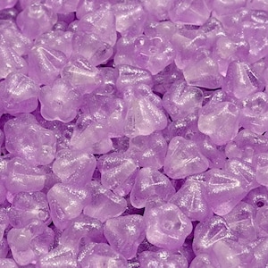 May include: A close-up shot of a pile of translucent, light purple beads. The beads have a faceted, irregular shape, with some resembling flowers or stars. The beads are scattered, creating a textured surface.