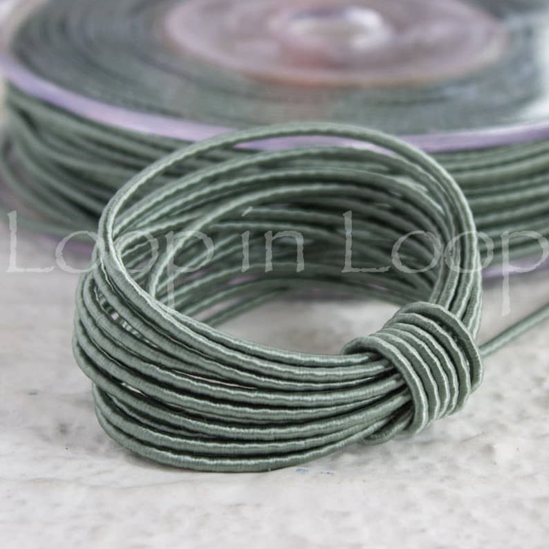 May include: A spool of sage green elastic cord. The cord is wound tightly and is ready to be used for crafting projects.