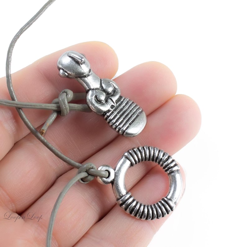 May include: A silver necklace with two pendants. One pendant is a stylized human figure and the other is a circle with a textured design. The necklace is made of a gray leather cord.