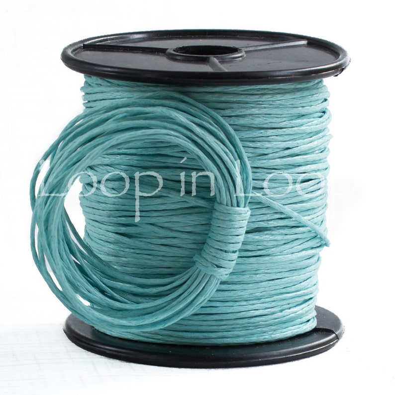 May include: A spool of light blue paper cord wrapped around a black plastic reel. The cord has a textured, fibrous appearance and is wound tightly. The text Loop in Loop is printed in white across the center of the blue cord spool.