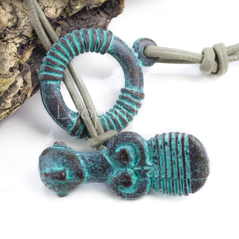 May include: A turquoise and brown leather necklace with two pendants. One pendant is a round ring with a textured surface. The other pendant is a stylized figure with a textured surface.