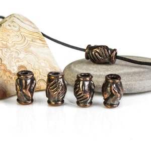 May include: Five copper-colored beads with a raised design of two hands clasped together. The beads are arranged in a row on a white surface.