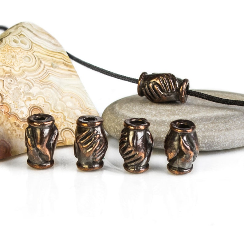 May include: Five copper-colored beads with a raised design of two hands clasped together. The beads are arranged in a row on a white surface.