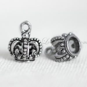 May include: Two silver crown-shaped charms. One charm is a detailed crown with a loop at the top. The other charm is a simple crown with a round opening.