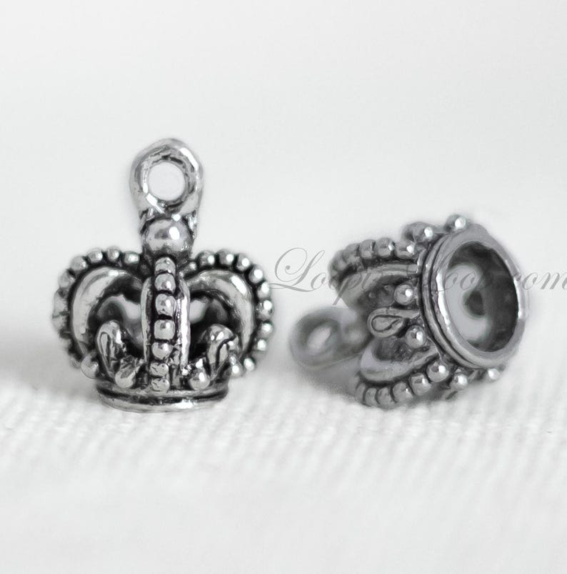 May include: Two silver crown-shaped charms. One charm is a detailed crown with a loop at the top. The other charm is a simple crown with a round opening.