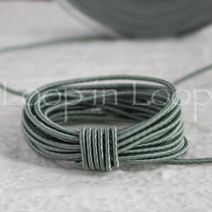 May include: A close-up of a coil of sage green elastic cord. The cord is thin and has a smooth, slightly shiny finish.