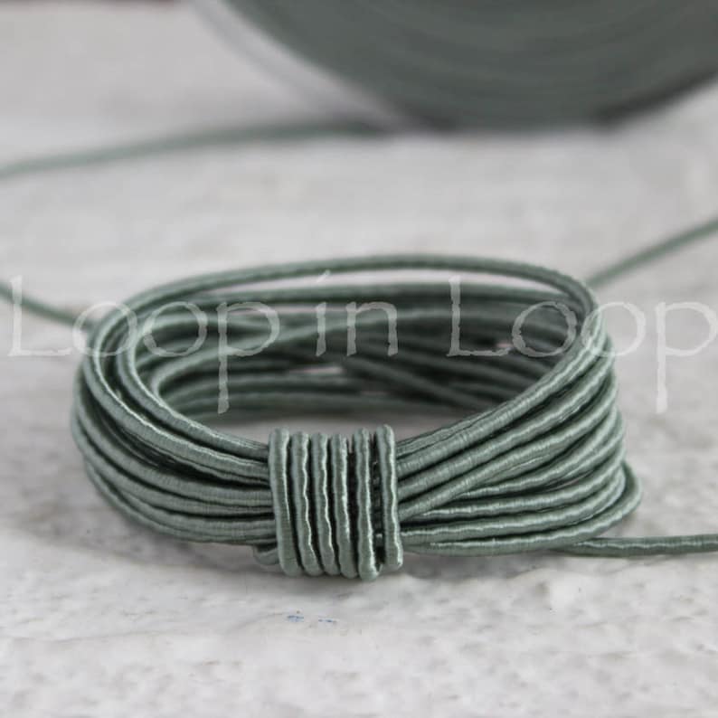 May include: A close-up of a coil of sage green elastic cord. The cord is thin and has a smooth, slightly shiny finish.