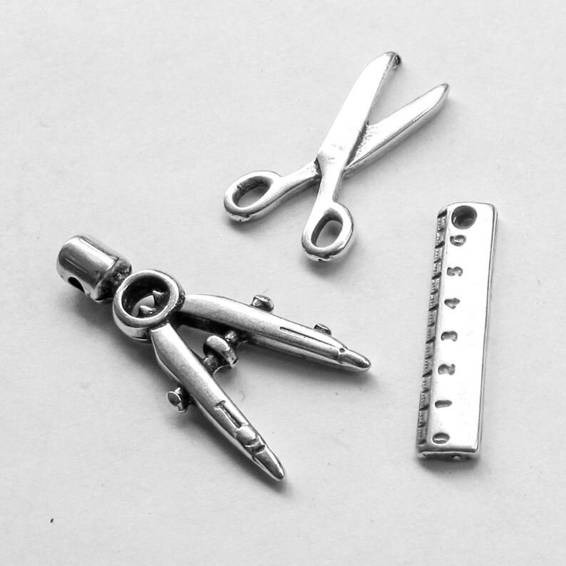 May include: Three silver charms in the shape of a compass, a pair of scissors, and a ruler. The ruler has numbers 0-5 printed on it.