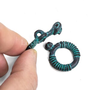 May include: Two antique-style metal clasps with a teal green patina. One clasp is a ring with a textured surface and the other is a hook with a decorative design.