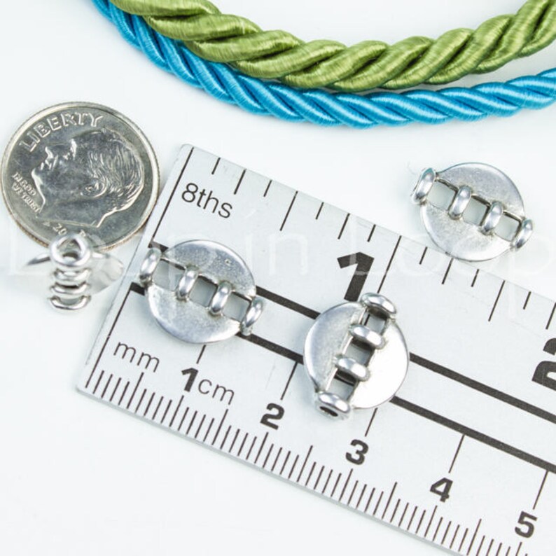 May include: Three silver metal cord ends with a ruler showing they are 1 cm in diameter. The cord ends have a design of four parallel lines.