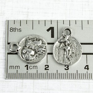 May include: Two silver-toned metal charms, each with a different design. One charm features a winged angel, while the other depicts a mythical creature. Both charms are approximately 1 cm in diameter and have a small loop for attaching to a necklace or bracelet.