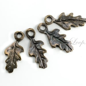 May include: Four bronze-colored oak leaf charms with a loop at the top for jewelry making or crafting. The leaves have detailed veining and a rustic, antique finish. The charms are arranged on a white background.