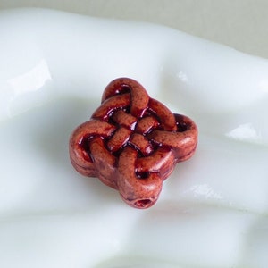 May include: A red clay bead with a Celtic knot design. The bead is square shaped and has a detailed, intricate design.