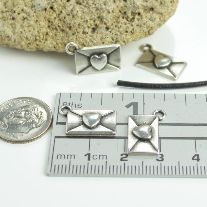 May include: Four silver-toned metal charms shaped like envelopes with a heart on the front. The charms are about 1 cm in size and are shown next to a ruler.