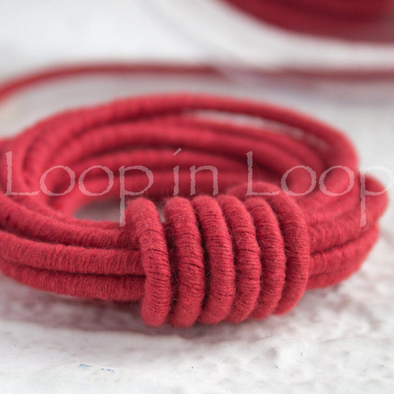 May include: Close-up of a red cotton cord, twisted and coiled into a circle. The cord is thick and soft, with a slightly textured surface.