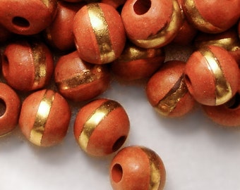 May include: A collection of terracotta beads with a gold stripe around the centre. The beads are round and have a hole through the centre for stringing.