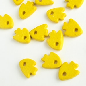 May include: A collection of yellow ceramic fish-shaped beads with a hole in the centre. The beads are scattered on a white surface.