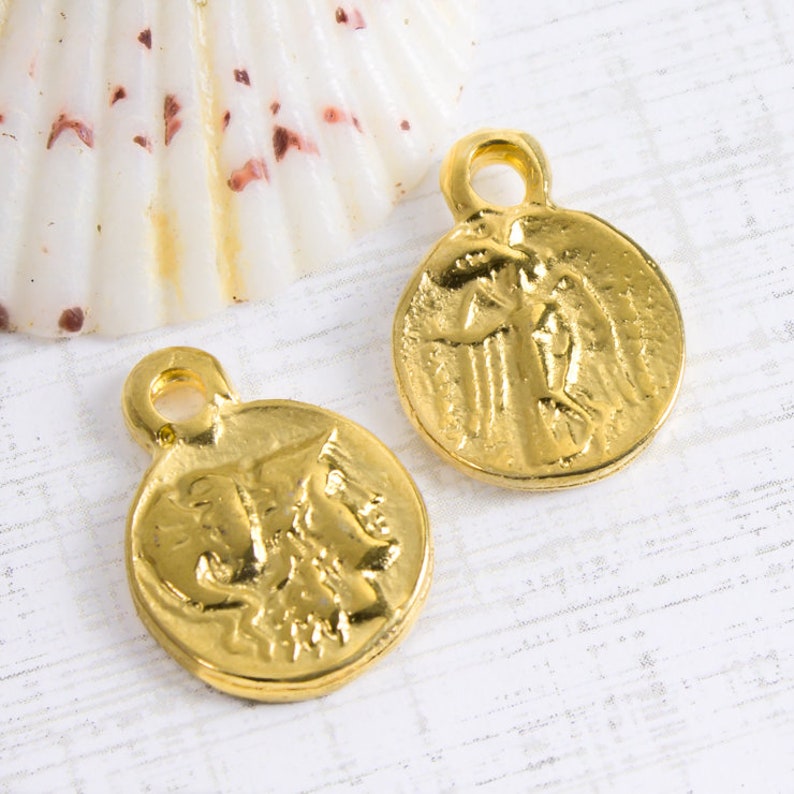May include: Two gold-colored metal charms in the shape of a round coin with a raised design of a winged figure. The charms have a small loop at the top for attaching to a necklace or bracelet.
