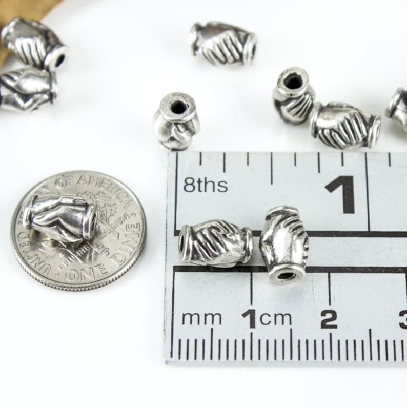 May include: Silver tone metal beads with a handshake design. The beads are approximately 1 cm in diameter and are shown next to a ruler for size reference.