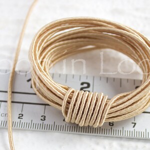 May include: A close-up of a spool of beige elastic cord. The cord is wound tightly around itself, creating a circular shape. The cord is about 2 inches in diameter.