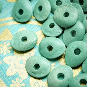 May include: A close-up of teal green wooden beads with a hole in the center. The beads are oval shaped and have a rough texture. The beads are scattered on a blue and gold patterned surface.