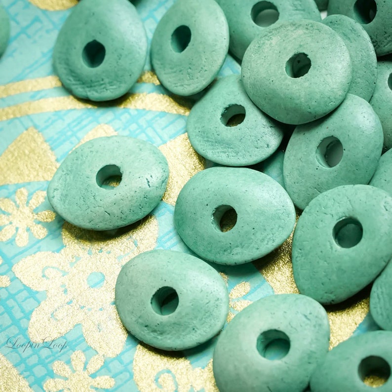 May include: A close-up of teal green wooden beads with a hole in the center. The beads are oval shaped and have a rough texture. The beads are scattered on a blue and gold patterned surface.