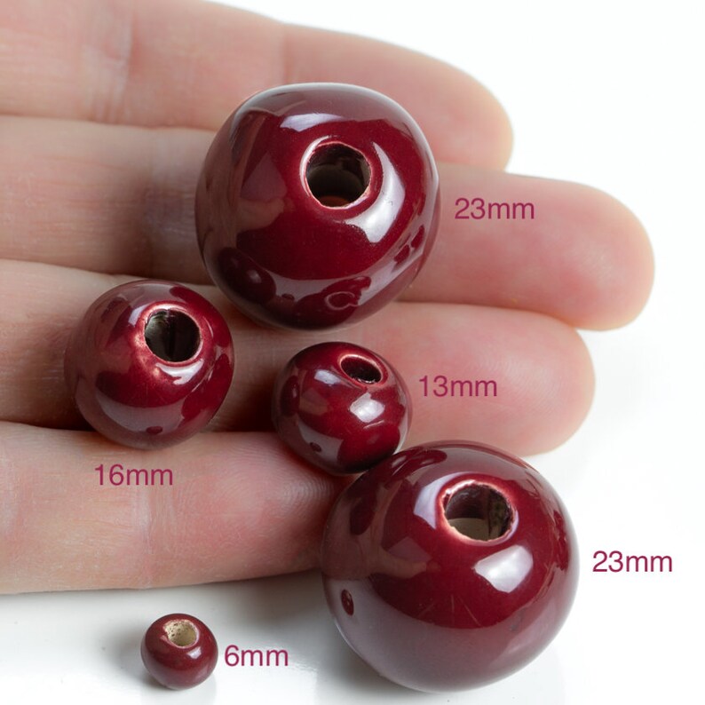 Op de afbeelding: Vijf rode keramische kralen in verschillende maten, met gaten in het midden. De kralen hebben een diameter van 6 mm, 13 mm, 16 mm en 23 mm.
