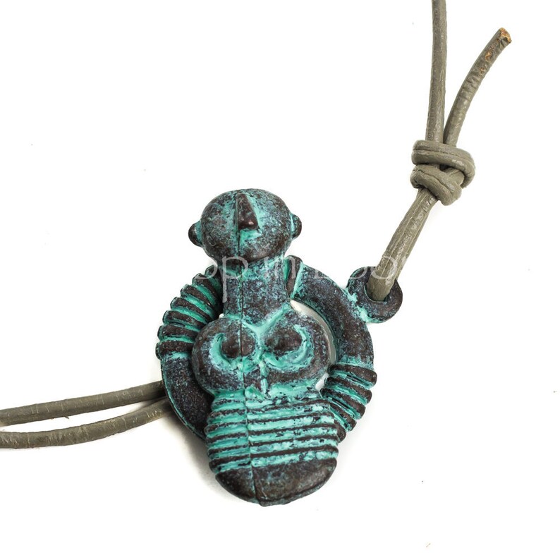 May include: A turquoise and brown metal pendant depicting a stylized female figure with a circular design. The pendant is attached to a gray leather cord.